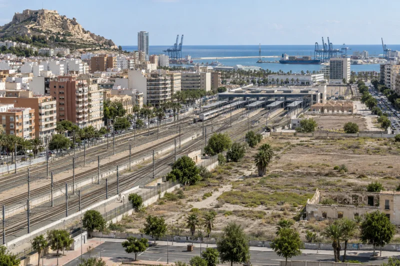 Vista de vías ferroviarias, edificios y grúas portuarias junto al Mediterráneo.