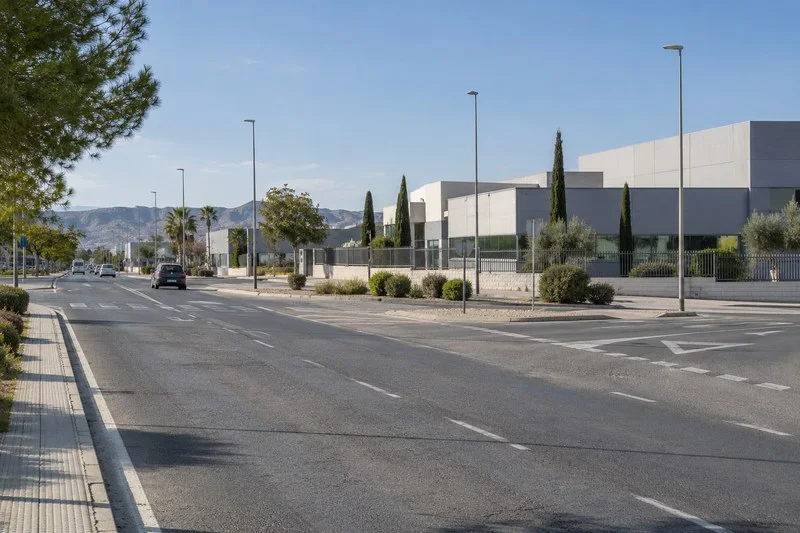 Una avenida asfaltada atraviesa un área de naves industriales; a un lado hay una acera arbolada y al fondo se ven montañas.