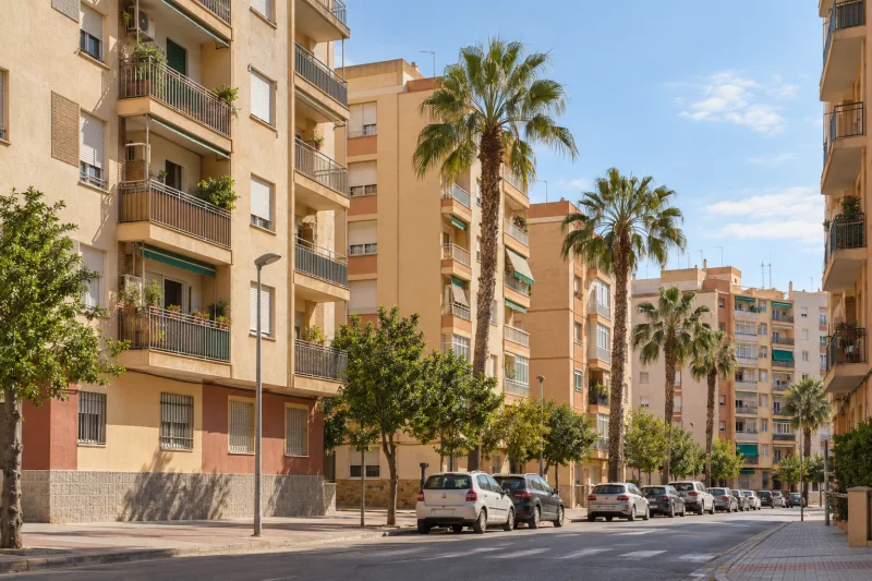 Vista de una calle residencial mediterránea con bloques de viviendas de varias plantas, balcones y palmeras.
