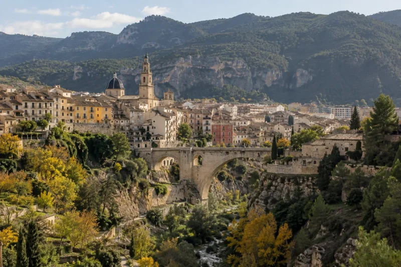 Vista panorámica de una ciudad entre montañas, con edificios y un puente de piedra sobre un barranco.