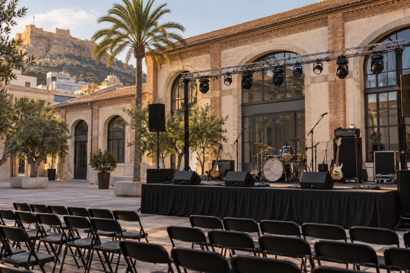 Escenario exterior preparado para un concierto, con batería, guitarras, amplificadores y sillas vacías frente a una fachada de ladrillo y piedra